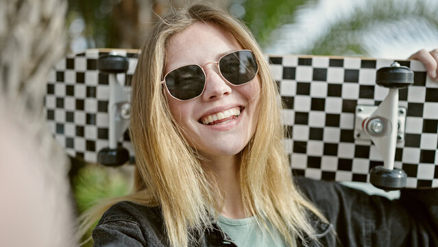 Young blonde woman holding skate make selfie by camera smiling at park