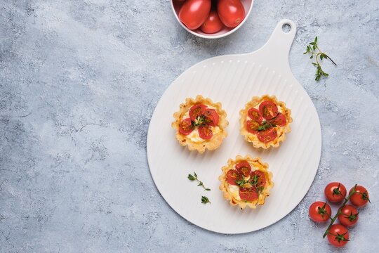 Unsweetened shortbread tartlets with feta cheese, cherry tomatoes and herbs on a white plate on a light concrete background. Unsweetened pastries,