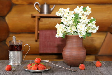 On a wooden table a metal plate of ripe strawberries is next to a bouquet of flowers in a pottery vase against a log wall on which a shelf  with utensils is hanging on it.