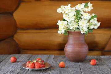 Obraz na płótnie Canvas On a wooden table a metal plate of ripe strawberries is next to a bouquet of flowers in a pottery jug against a log wall.