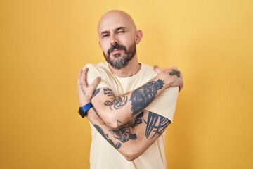 Hispanic man with tattoos standing over yellow background hugging oneself happy and positive,...