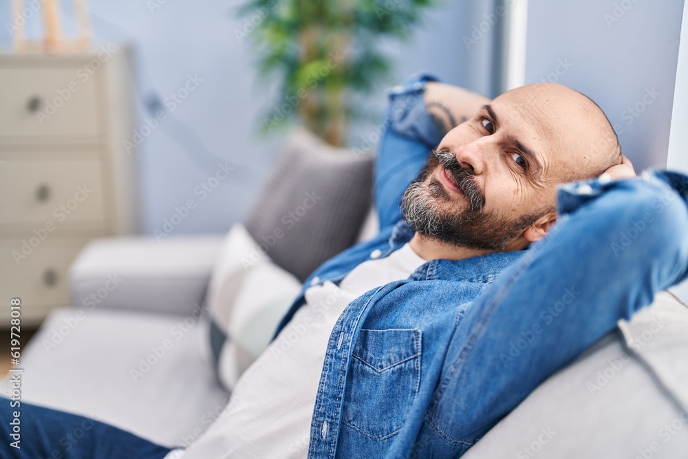 Poster young bald man relaxed with hands on head sitting on sofa at home