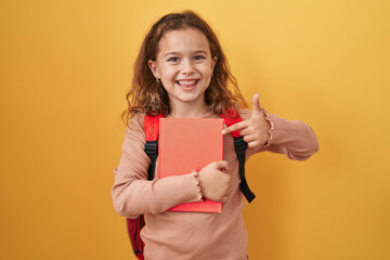 Fototapeta na wymiar Little caucasian girl wearing student backpack and holding book smiling happy pointing with hand and finger