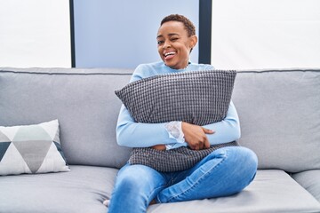 African american woman sitting on the sofa at home hugging pillow winking looking at the camera...