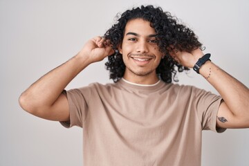 Hispanic man with curly hair standing over white background smiling pulling ears with fingers,...