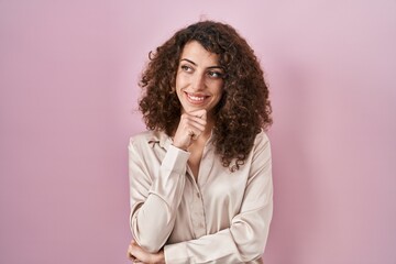 Fototapeta premium Hispanic woman with curly hair standing over pink background with hand on chin thinking about question, pensive expression. smiling and thoughtful face. doubt concept.