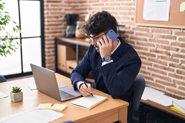 Naklejka premium Young hispanic man business worker talking on smartphone write on notebook at office