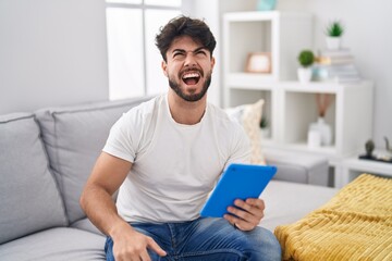 Hispanic man with beard using touchpad sitting on the sofa angry and mad screaming frustrated and furious, shouting with anger. rage and aggressive concept.