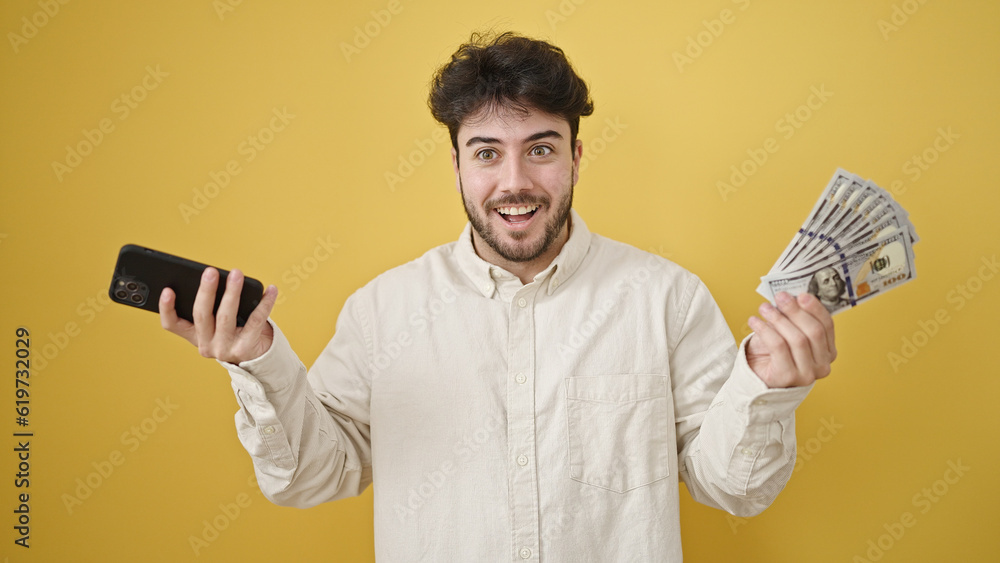 Wall mural young hispanic man smiling holding dollars and smartphone over isolated yellow background