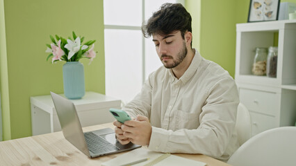 Young hispanic man using laptop and smartphone sitting on table at dinning room