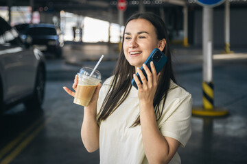 A young woman with coffee talking on the phone in the parking lot.
