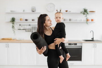 Smiling brunette woman holding barefoot infant girl and rubber mat in arms on kitchen background....