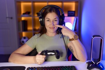 Beautiful brunette woman playing video games wearing headphones pointing with hand finger to face...