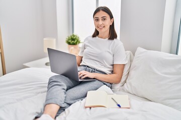 Young hispanic woman sitting on bed using laptop studying at bedroom