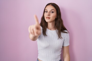 Young hispanic girl standing over pink background pointing with finger up and angry expression, showing no gesture