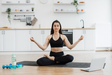 Relaxed slim woman in black activewear meditating in lotus pose with hands in gyan mudra to music from laptop. Attractive lady enjoying calming tunes during yoga practice in modern apartment.