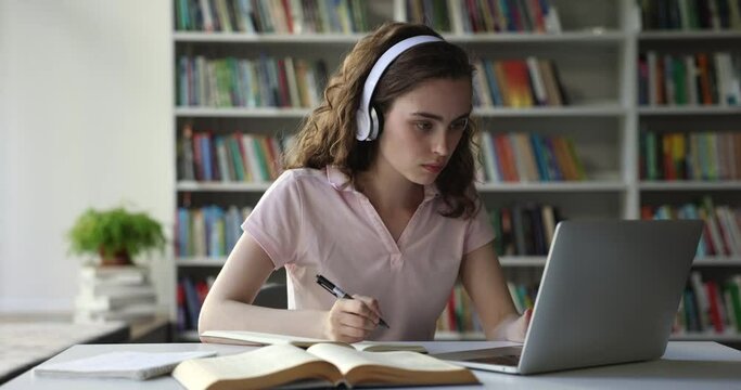 Serious pretty college student girl in wireless headphones watching video lesson, class, listening online learning course on laptop in library, writing notes at table, getting positive, happy