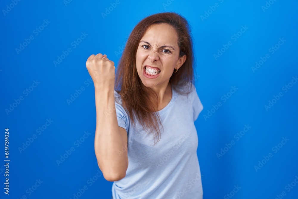 Poster Brunette woman standing over blue background angry and mad raising fist frustrated and furious while shouting with anger. rage and aggressive concept.