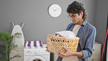 Young hispanic man holding basket smelling clothes at laundry room