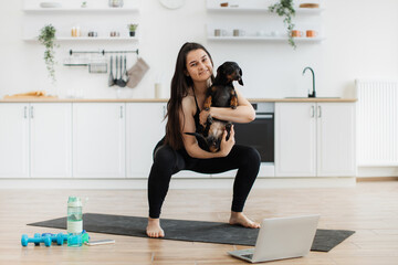 Fototapeta na wymiar Beautiful woman in sportswear cuddling Dachshund while doing squats on rubber mat in modern apartment. Athletic person strengthening balance and coordination together with buddy pet in arms at home.
