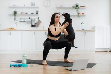 Young slim female in athletic wear holding black dog in arms while crouching on yoga mat in kitchen...