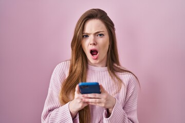 Young caucasian woman using smartphone typing message angry and mad screaming frustrated and...