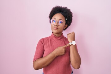 Beautiful african woman with curly hair standing over pink background in hurry pointing to watch...