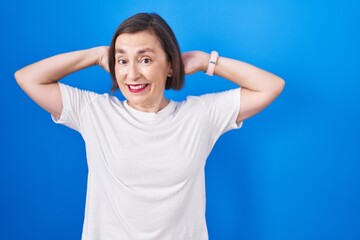 Fototapeta premium Middle age hispanic woman standing over blue background relaxing and stretching, arms and hands behind head and neck smiling happy