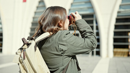 Mature hispanic woman with grey hair tourist wearing backpack taking pictures with vintage camera at street