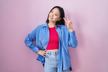 Young asian woman standing over pink background smiling looking to the camera showing fingers doing...