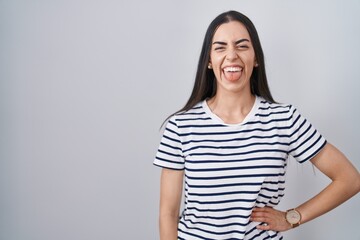 Fototapeta premium Young brunette woman wearing striped t shirt sticking tongue out happy with funny expression. emotion concept.