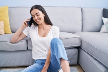 Young hispanic woman talking on the smartphone sitting on floor at home