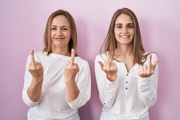 Middle age mother and young daughter standing over pink background showing middle finger doing fuck...