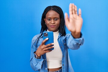 African woman with braids using smartphone typing message with open hand doing stop sign with...