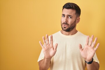 Handsome hispanic man standing over yellow background moving away hands palms showing refusal and...