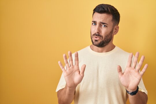 Handsome hispanic man standing over yellow background moving away hands palms showing refusal and denial with afraid and disgusting expression. stop and forbidden.