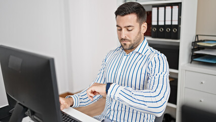 Young hispanic man business worker using computer looking watch at office