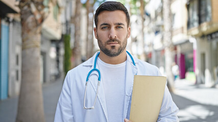 Young hispanic man doctor holding medical report standing with relaxed expression at street