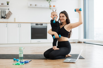 Fototapeta na wymiar Full length portrait of sporty lady and cute little child with dumbbells sitting on yoga mat in modern kitchen. Happy mother focusing on healthy routine together with small daughter on weekend.