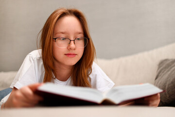 The girl is reading a thick book lying on the sofa.