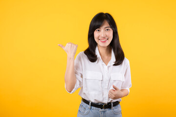 Happy young asian woman wearing white shirt and denim jean plants pointing thumb finger away on copy space isolated on yellow background. helps pick best choice, recommends product concept.