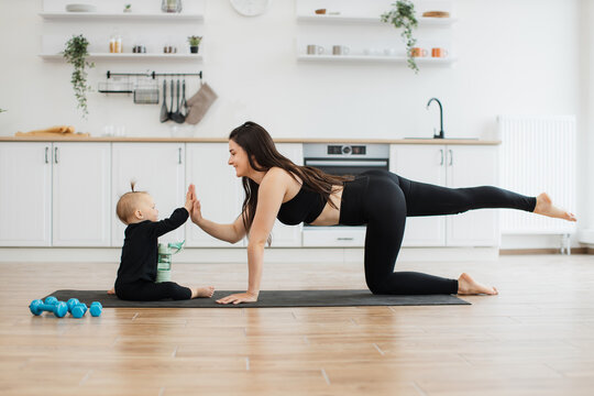 Funny small kid in black outfit responding to lady's high-five gesture while sitting near parent on floor. Affectionate mother establishing strong connection between her and child during home workout.