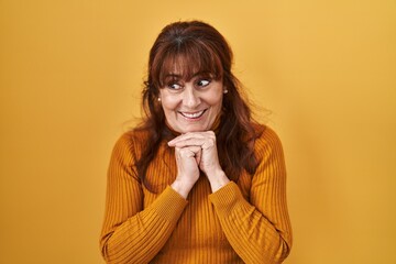 Middle age hispanic woman standing over yellow background laughing nervous and excited with hands on chin looking to the side