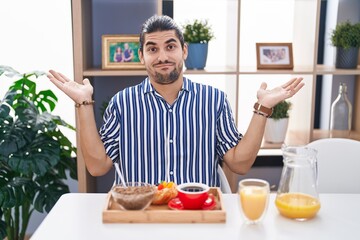 Hispanic man with long hair sitting on the table having breakfast clueless and confused expression with arms and hands raised. doubt concept.