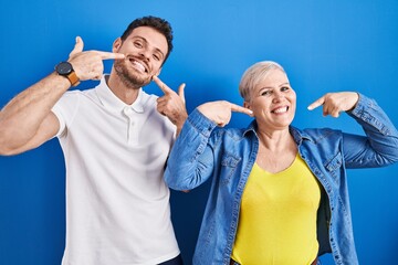 Young brazilian mother and son standing over blue background smiling cheerful showing and pointing with fingers teeth and mouth. dental health concept.