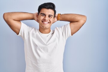 Hispanic man standing over blue background relaxing and stretching, arms and hands behind head and...