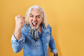 Middle age man with grey hair standing over yellow background angry and mad raising fist frustrated...