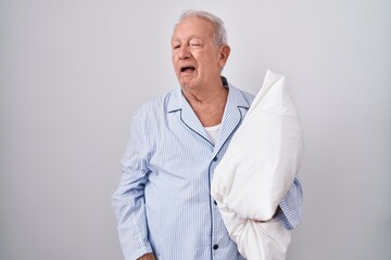 Senior man with grey hair wearing pijama hugging pillow winking looking at the camera with sexy...