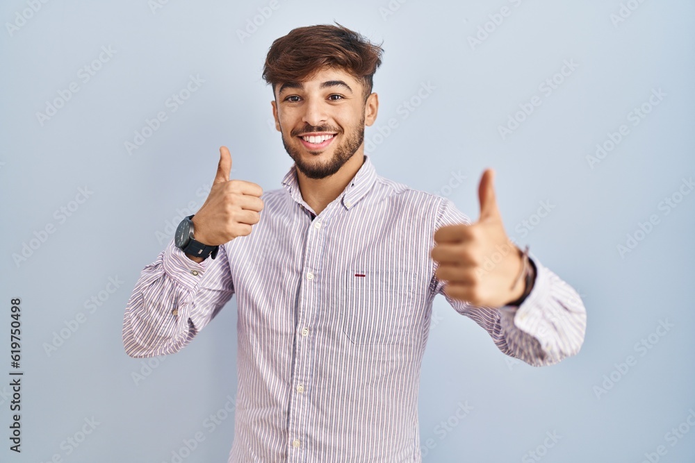 Wall mural arab man with beard standing over blue background success sign doing positive gesture with hand, thu