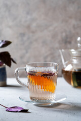 Healthy purple basil tea in a glass mug with slice of orange, teapot and fresh leaves backside. Grey concrete background, vertical banner with space for text
